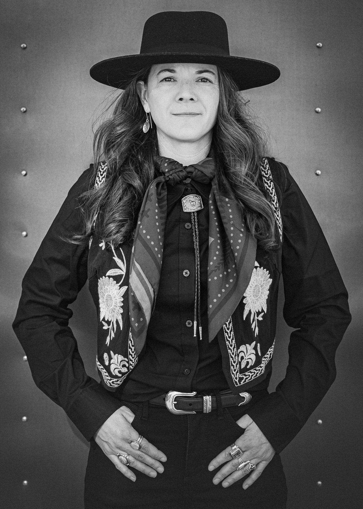 A black and white portrait of a woman wearing a wide-brimmed cowboy hat, a black western shirt, a patterned embroidered scarf, a bolo tie, and a belt, posed against a textured metal background.