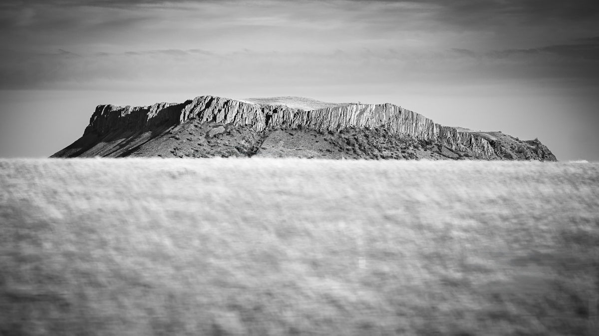Black and white landscape photograph of a rugged rocky cliff or mesa rising above a textured field, with a hazy sky above.