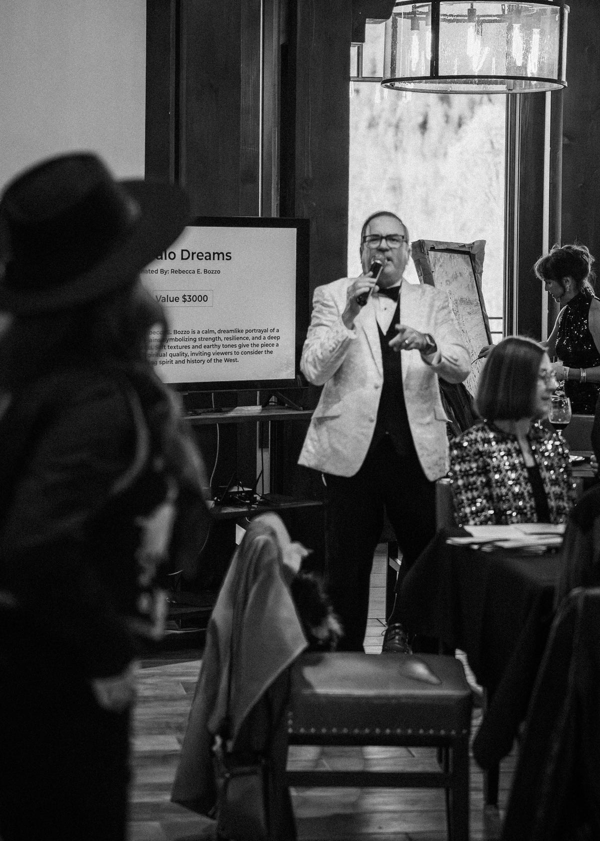 Male auctioneer in a suit standing at a podium with a screen behind him in a formal setting.
