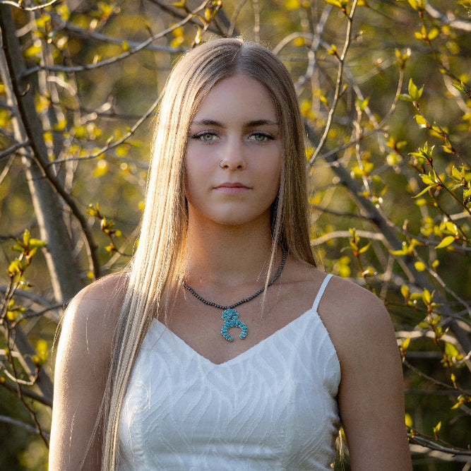 Woman in a white dress standing in front of trees with a necklace